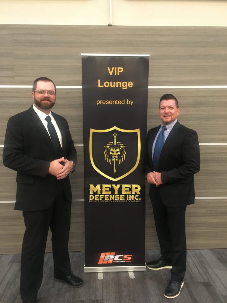 Two men in formal attire standing beside a banner that reads "VIP Lounge presented by Mayer Security Services," featuring a logo with a lion and a sword, emphasizing professional security and defense services.